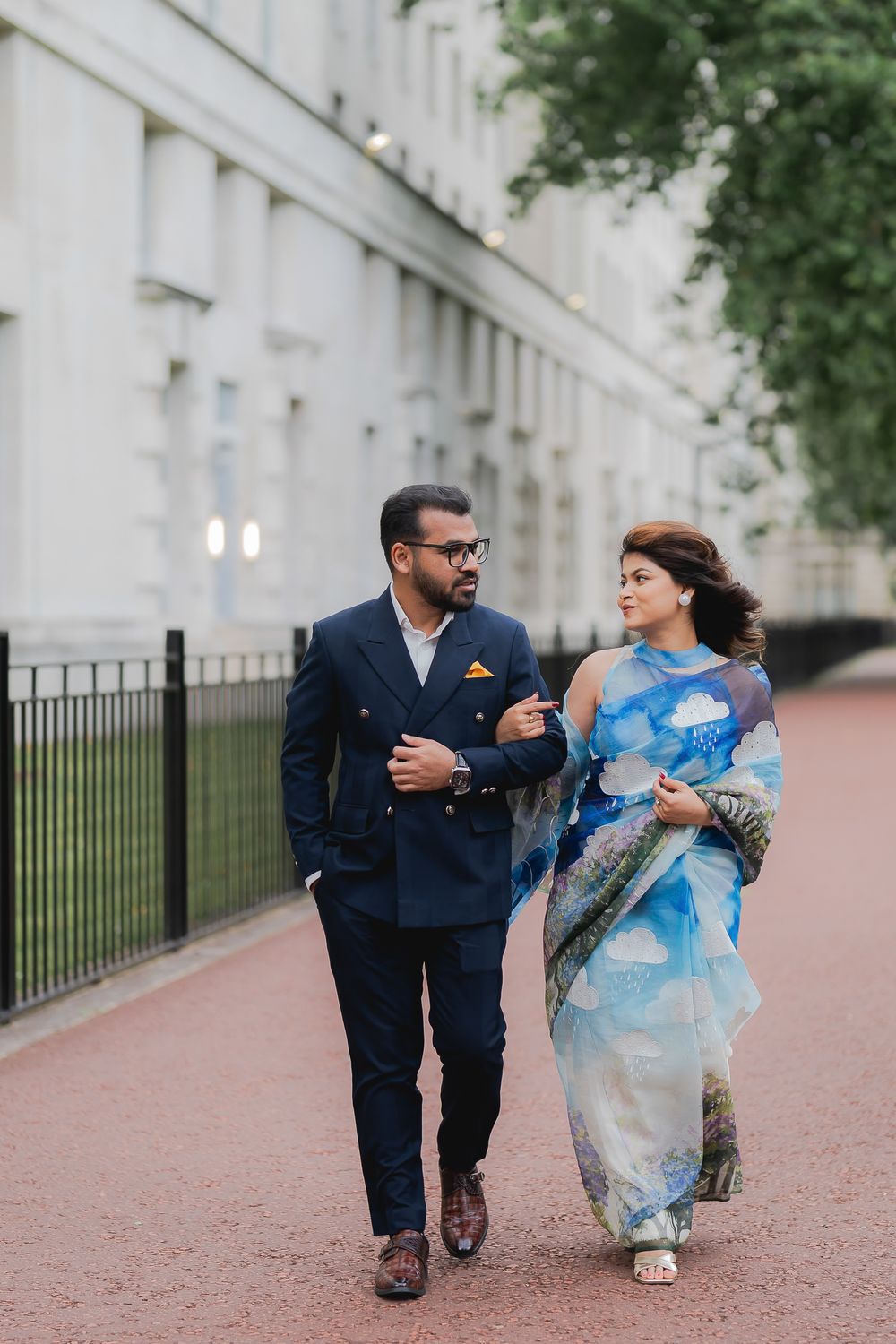 Indian couple walking together, man in navy suit and woman in blue printed saree, strolling along elegant building path.