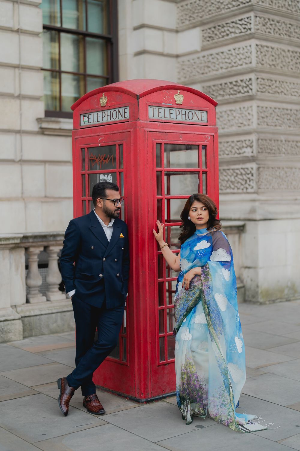 Couple in traditional Indian attire poses by iconic red London telephone box, blending cultures elegantly.