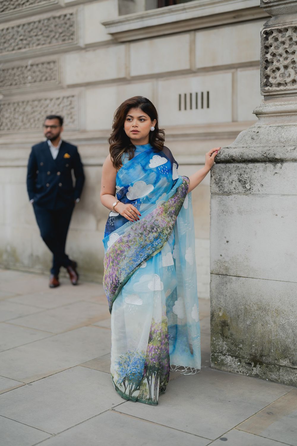 Woman in blue floral saree with sky print poses by stone column, man in navy suit stands in background.