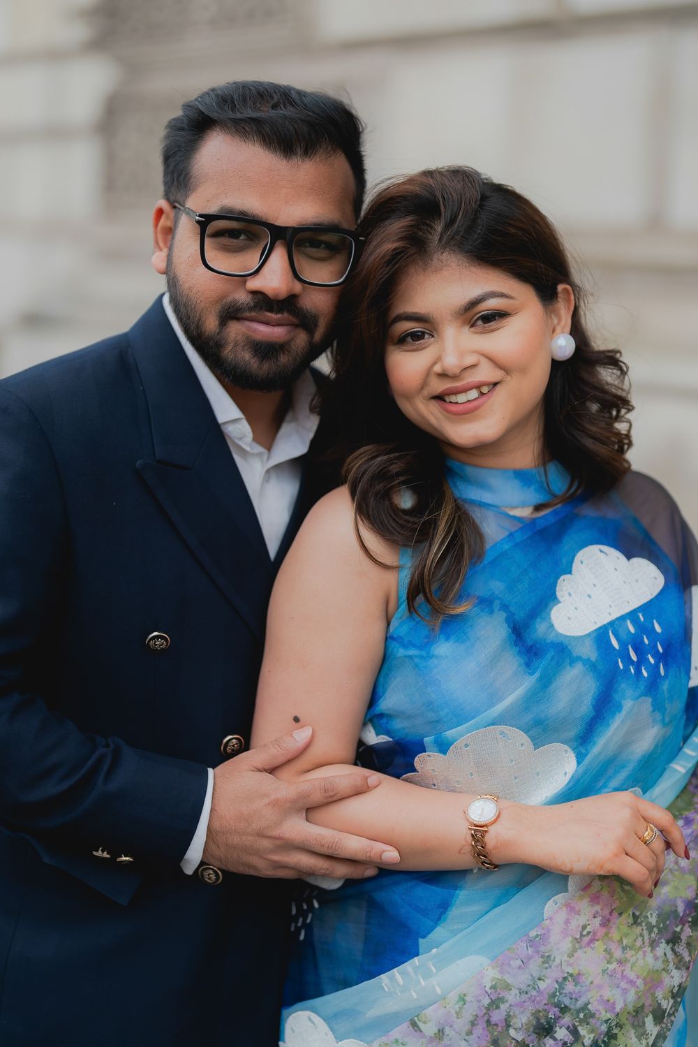 A smiling couple poses together, man in navy blazer and glasses embracing woman in blue cloud-print dress.
