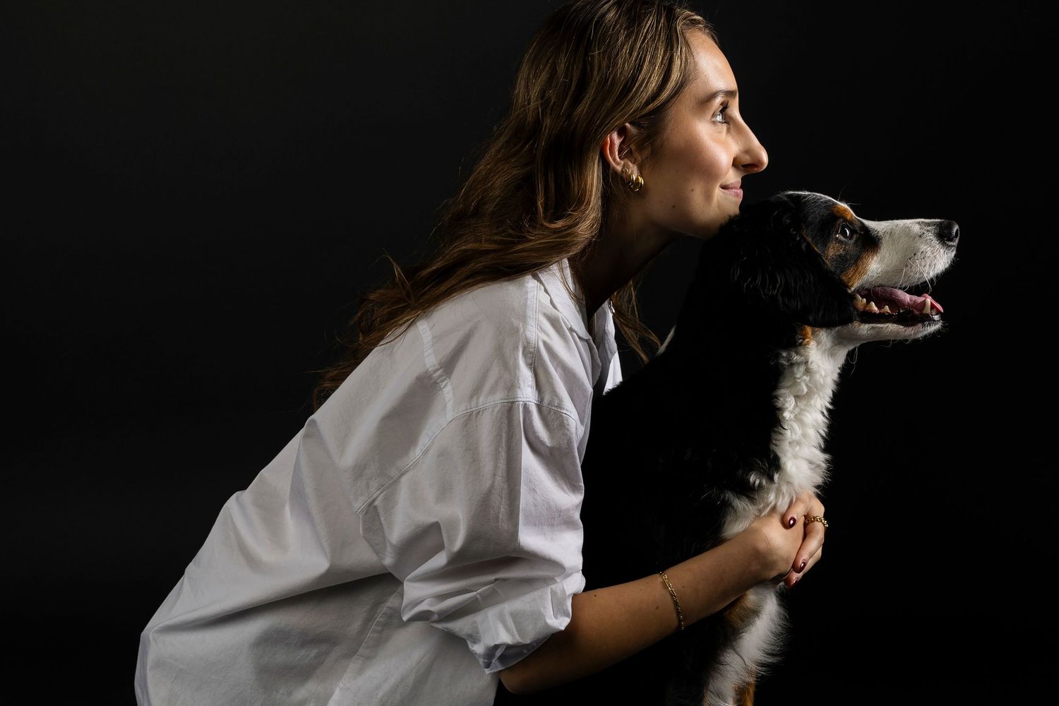 A woman in a white blouse holds a black tri australian cattle dog against a dark background, both looking to the side.