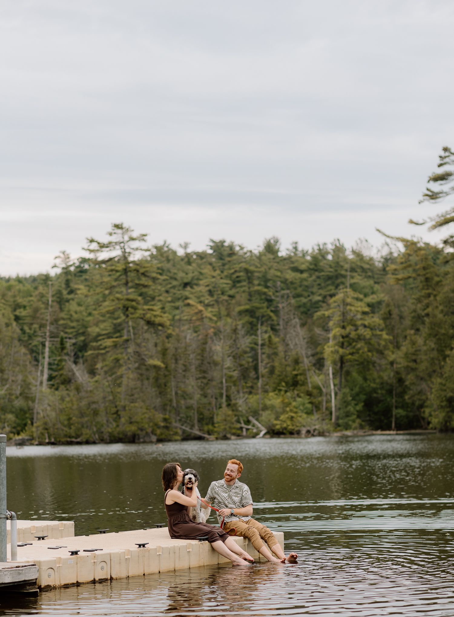 Couple sitting on a wooden dock with feet in the water, surrounded by a serene lake and lush forest backdrop.