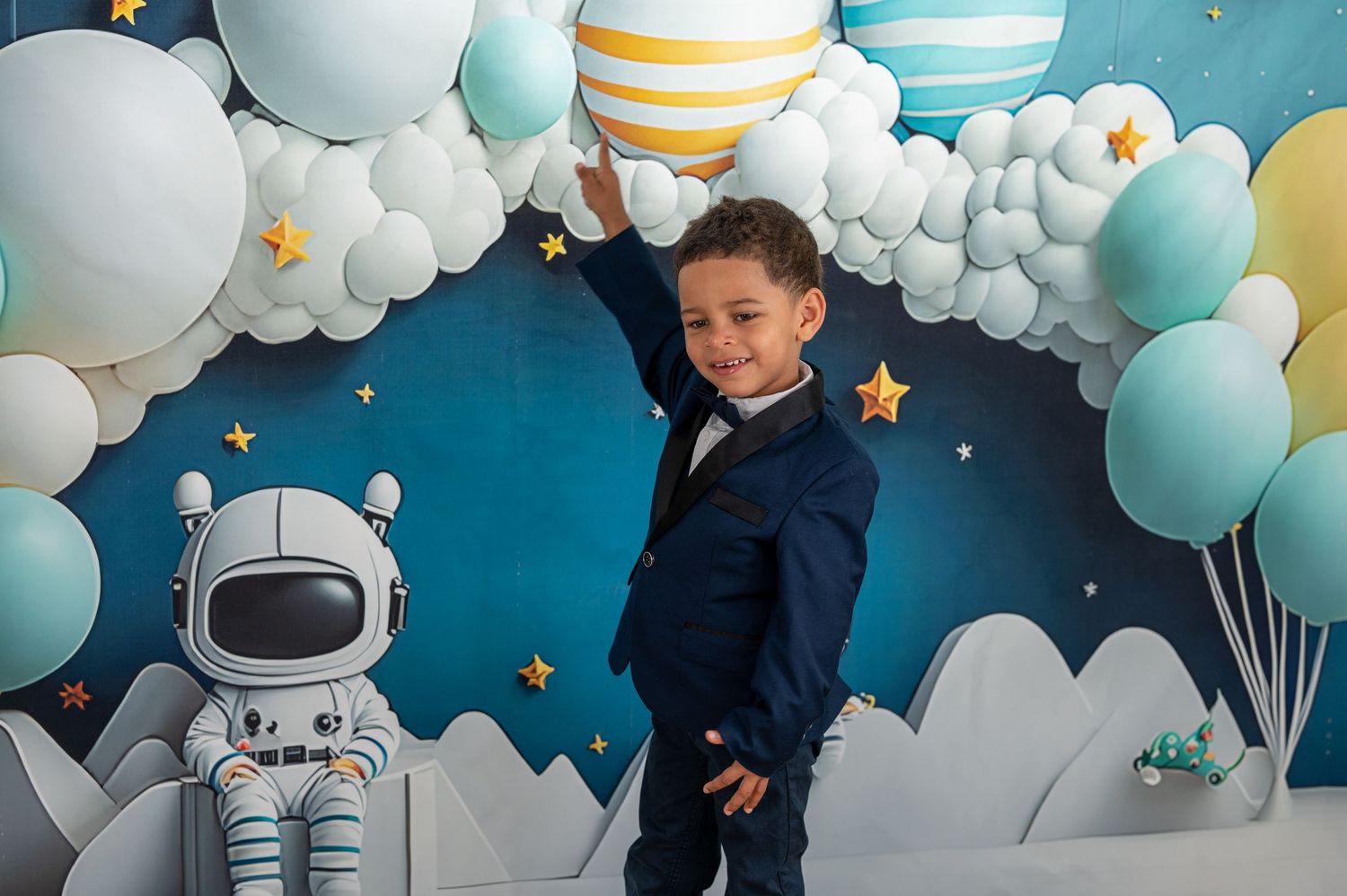 Un jeune garçon en costume bleu marine pose devant une toile de fond sur le thème de l'espace avec des ballons, des planètes et une figurine