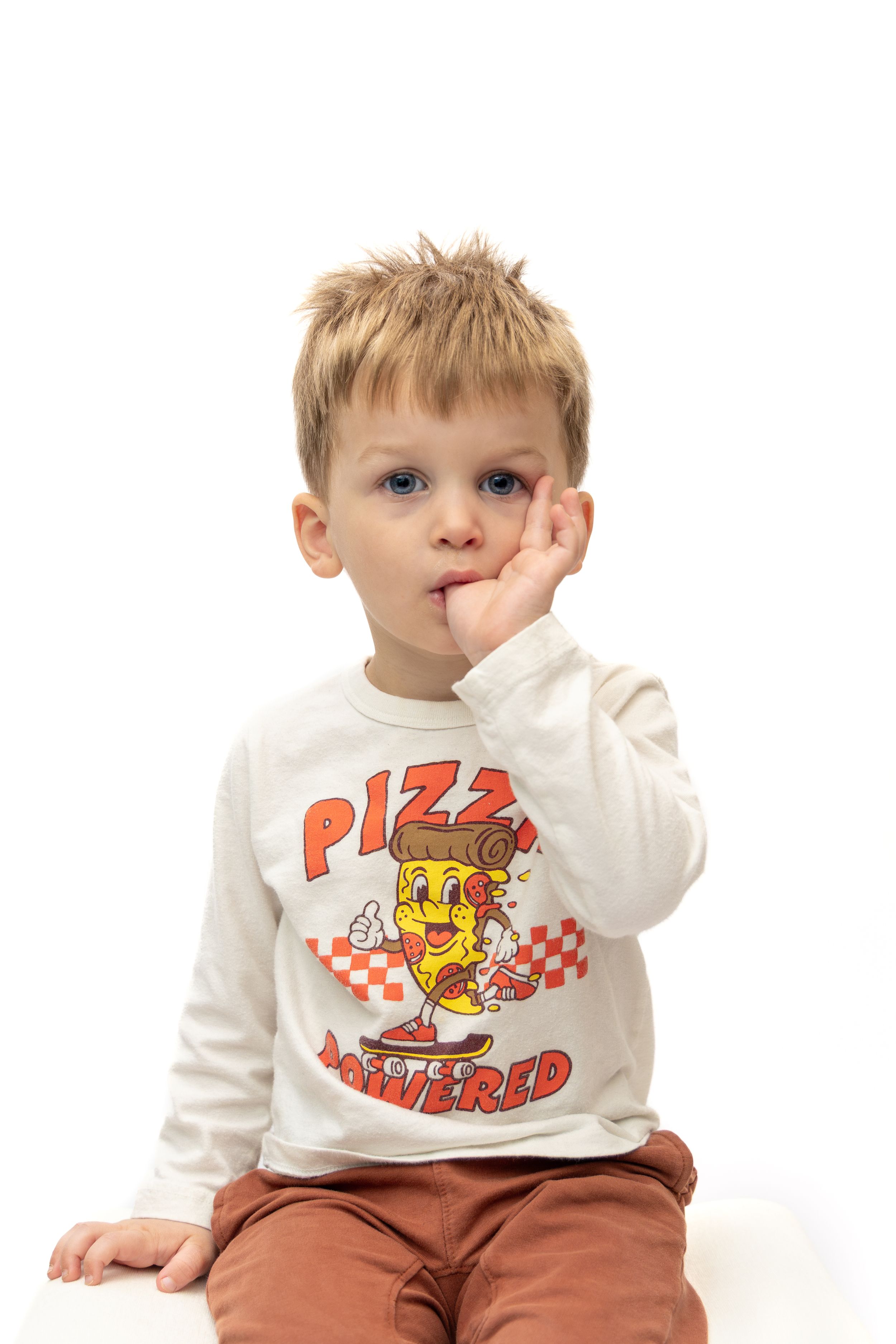 Cute toddler boy with blonde hair wearing a white Pizza Powered graphic long sleeve shirt and brown pants.