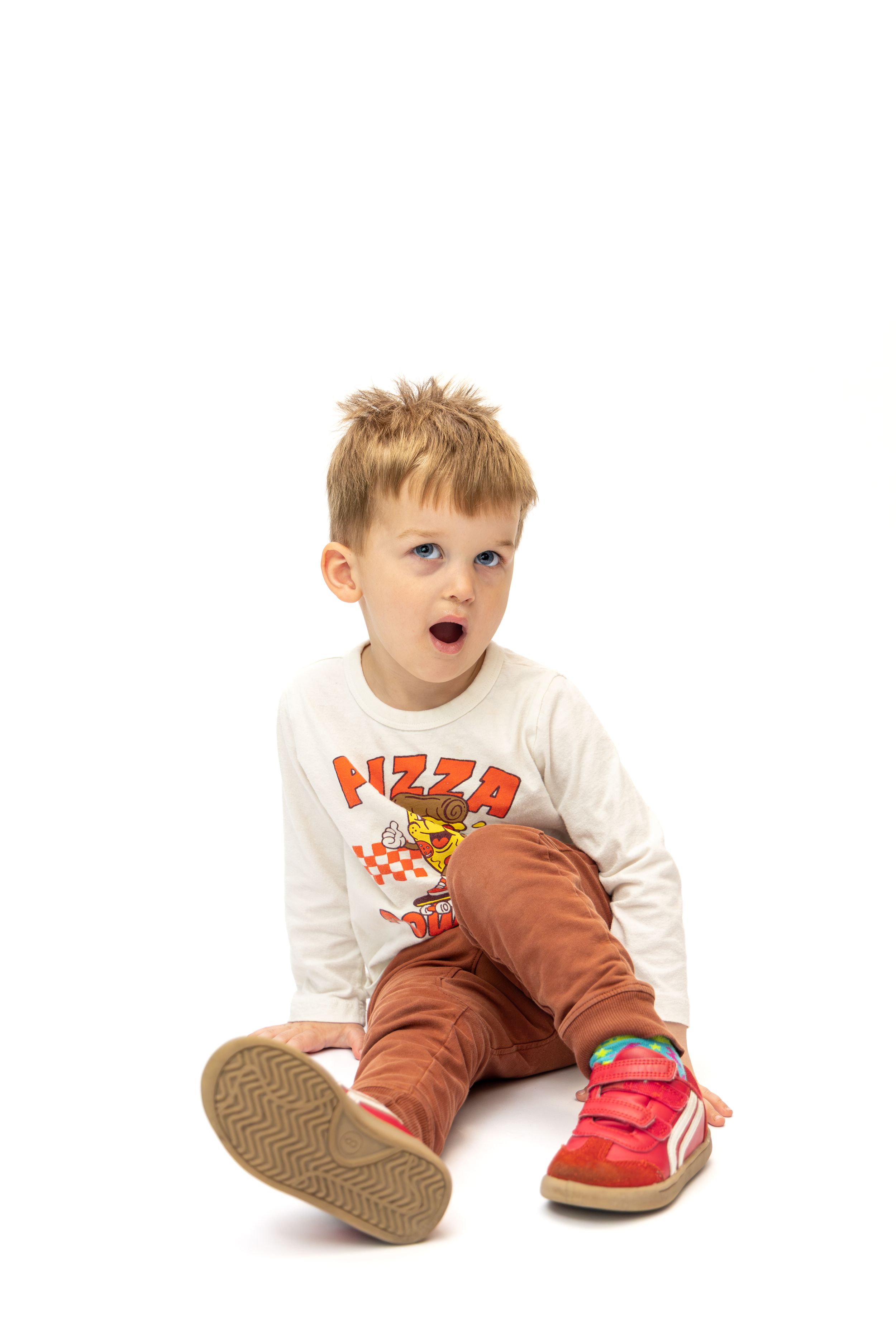 Cute toddler boy in a white pizza shirt, brown pants, and red sneakers sitting on a white background.