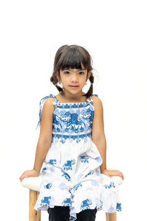 Young girl in blue and white elephant print dress sitting on wooden chair against white background.