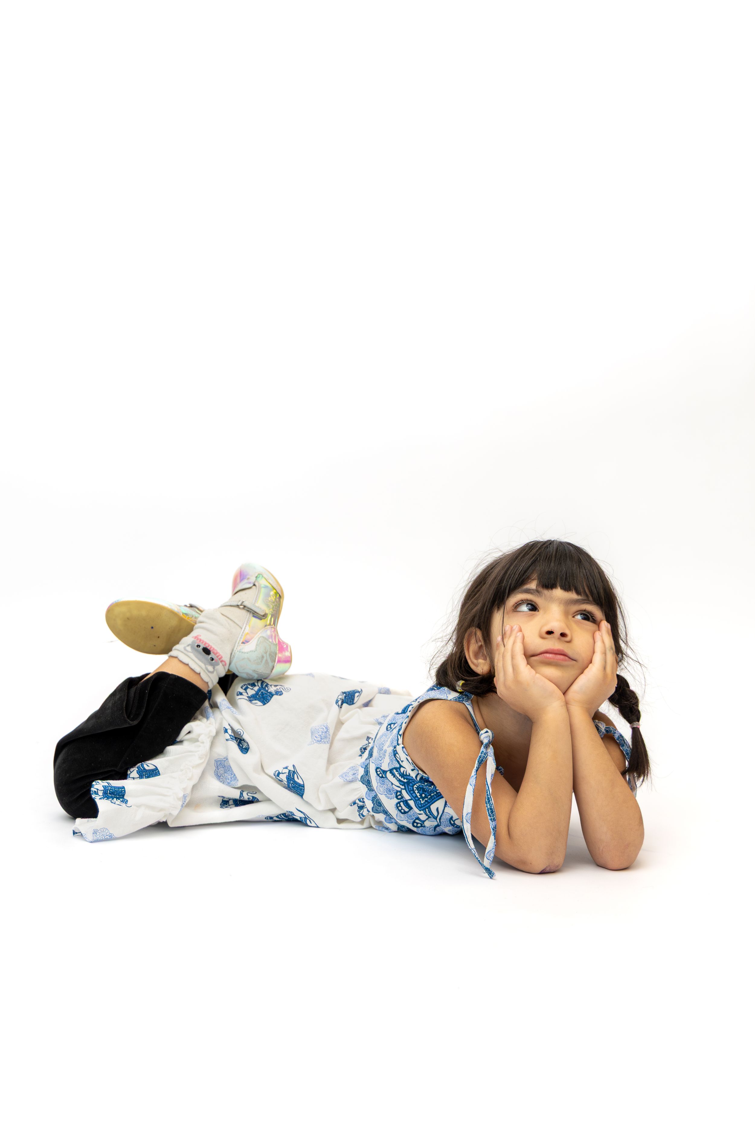 A young girl in a floral dress lies on the floor, resting her chin on her hands, gazing upward thoughtfully.