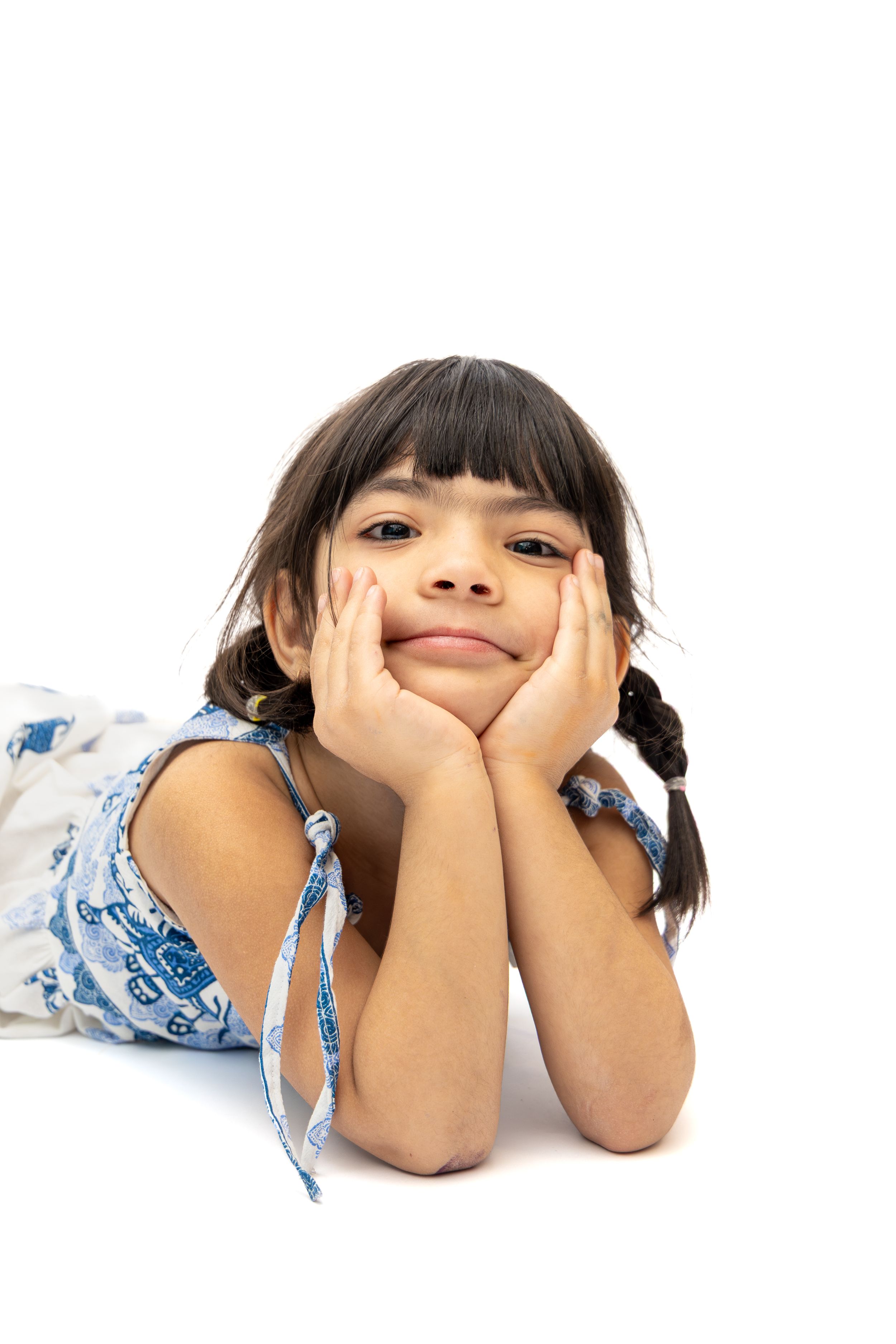 Young girl with braids resting chin on hands, lying down in blue dress, smiling at camera on white background.