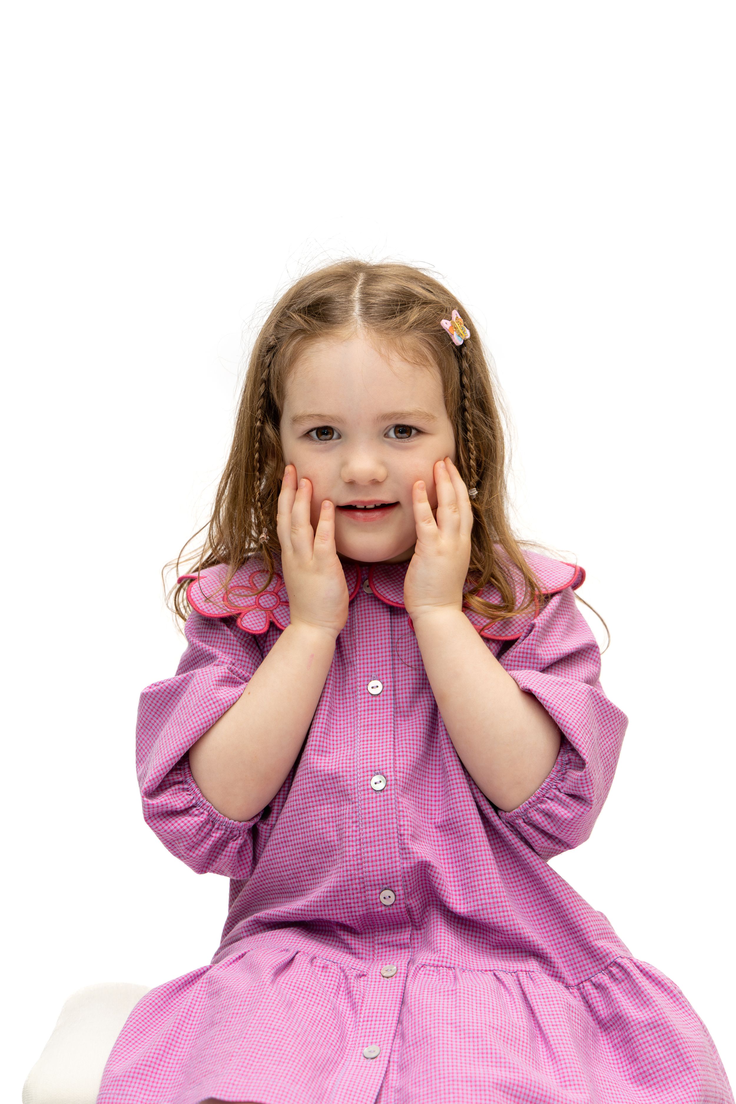 A cute little girl in a pink dress sits with hands on cheeks, looking surprised against a white background.