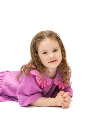 A young girl in a pink dress with floral embroidery lies on a white background, smiling sweetly at the camera.