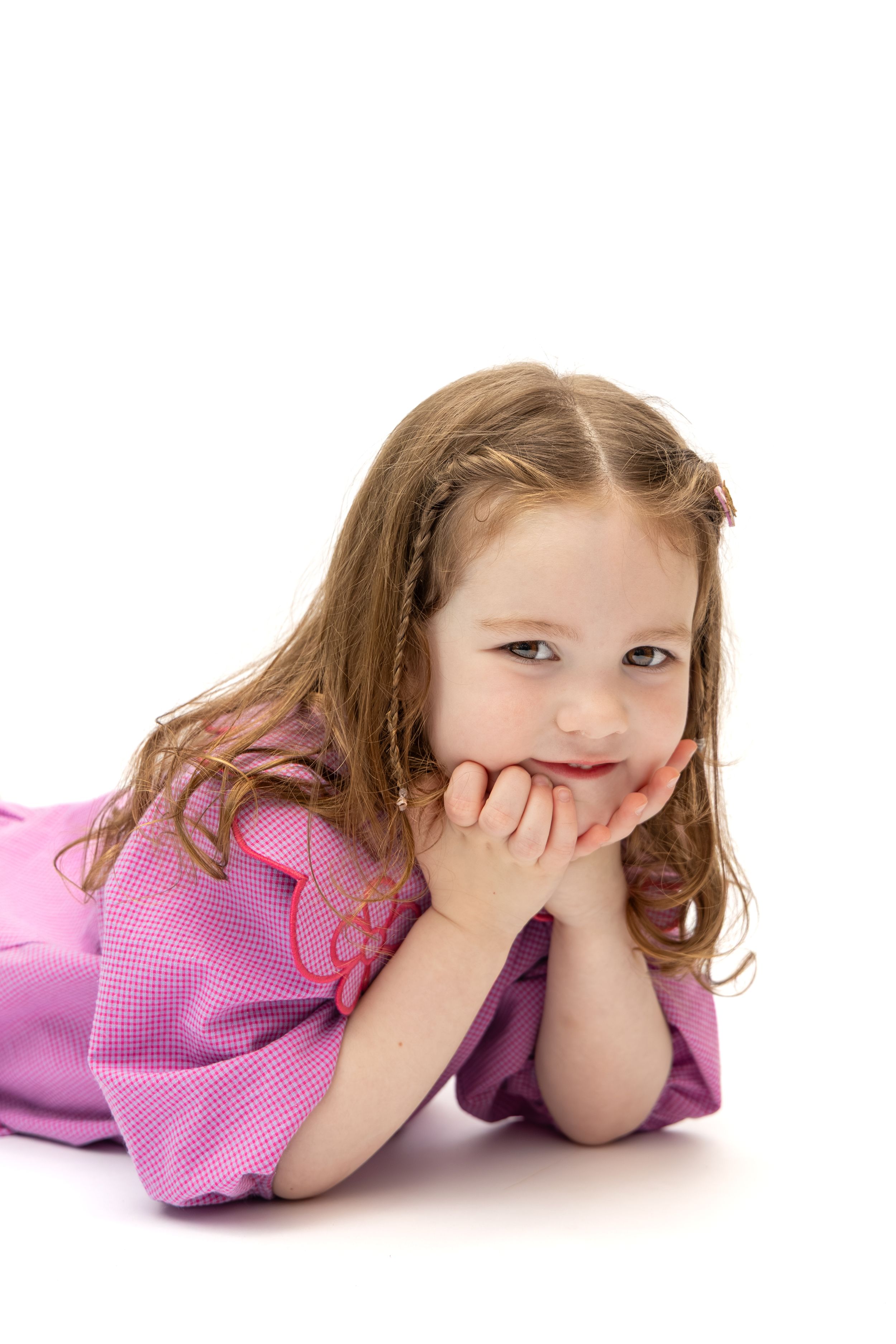 A young girl in a pink dress lying down with her chin resting on her hands, smiling sweetly against a white background.