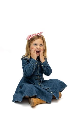 A surprised little girl in a denim dress and red bow sits on the floor with hands on cheeks against white background.