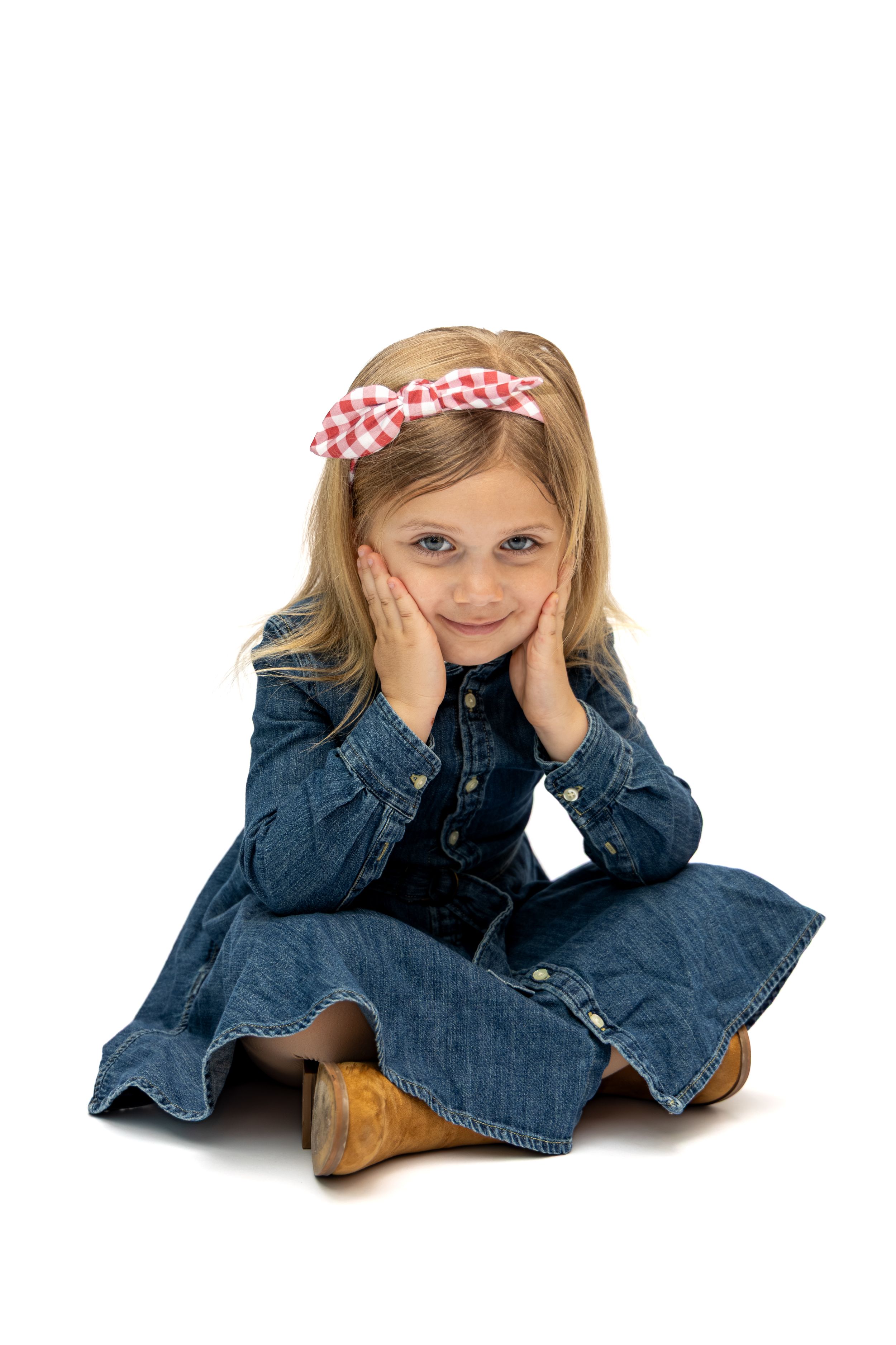 Young girl in denim dress and red checkered bow sits cross-legged, hands on cheeks, smiling on white background.