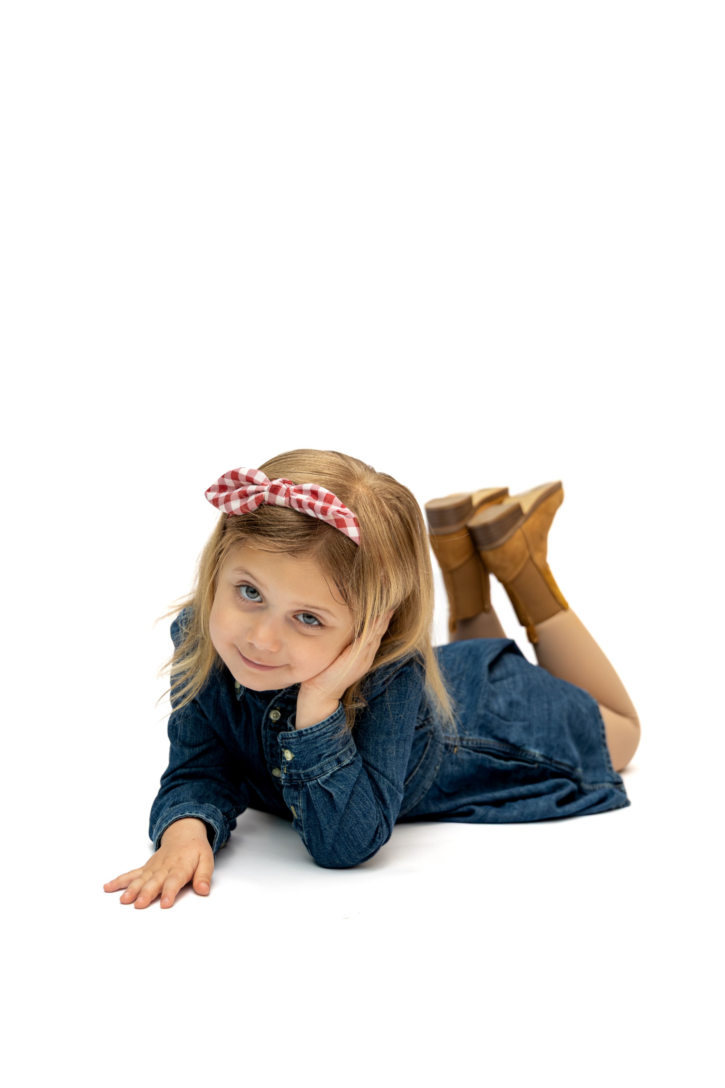 Young girl in denim shirt and red bow headband lying on floor smiling, wearing brown boots on white background.