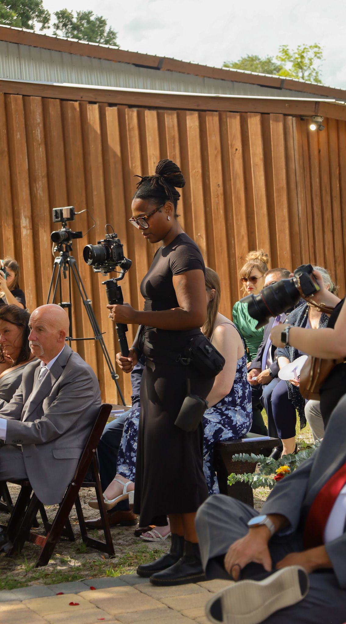 A Black female photographer in a black dress captures moments at an outdoor wedding ceremony with guests seated on chairs.