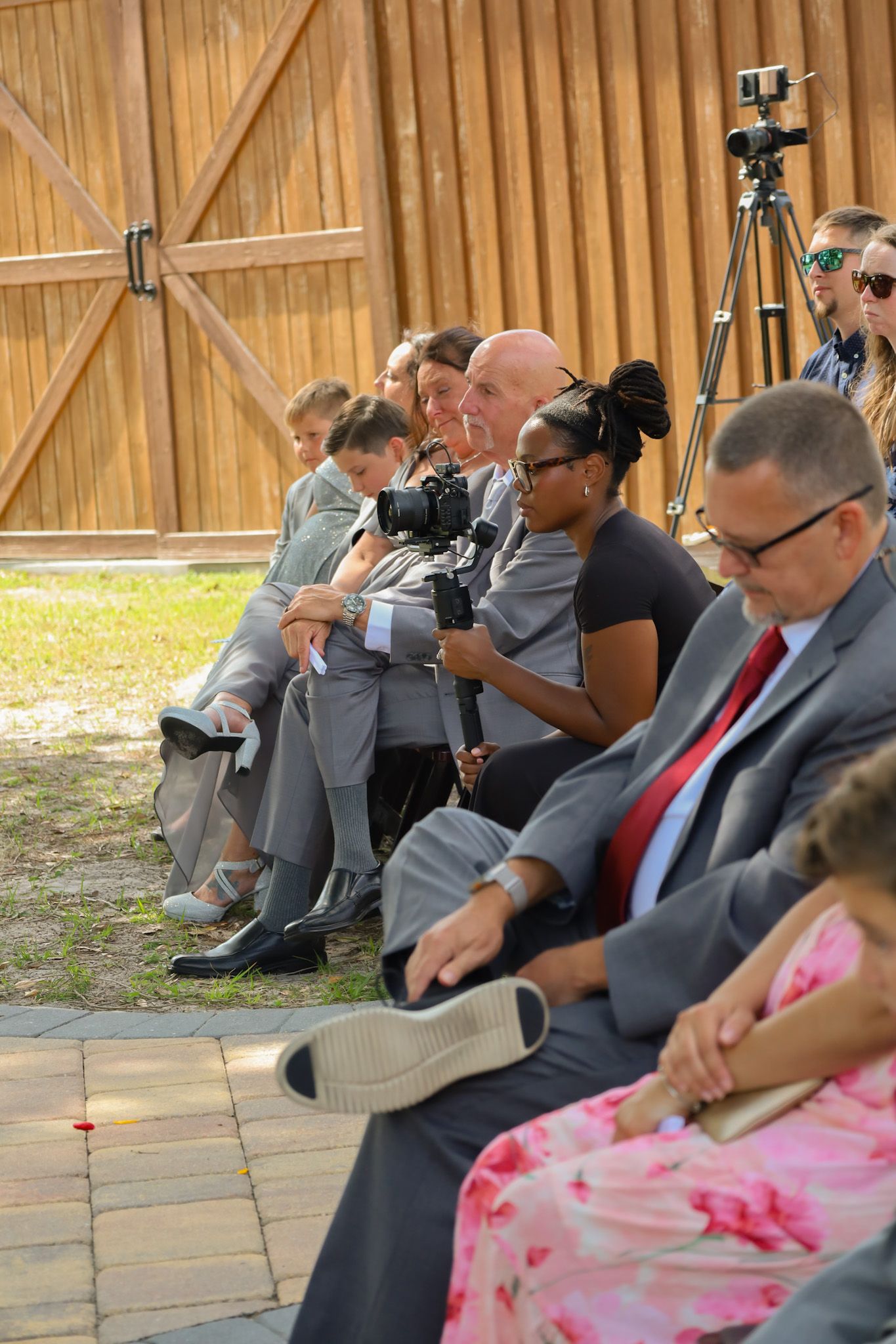 Guests seated outdoors at a wedding ceremony, with a photographer holding a camera and tripods visible near a wooden barn.