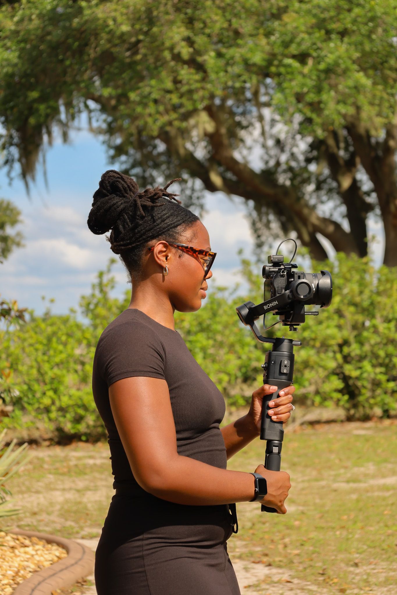 A Black woman with dreadlocks holds a camera gimbal stabilizer outdoors, wearing a brown top and sunglasses.