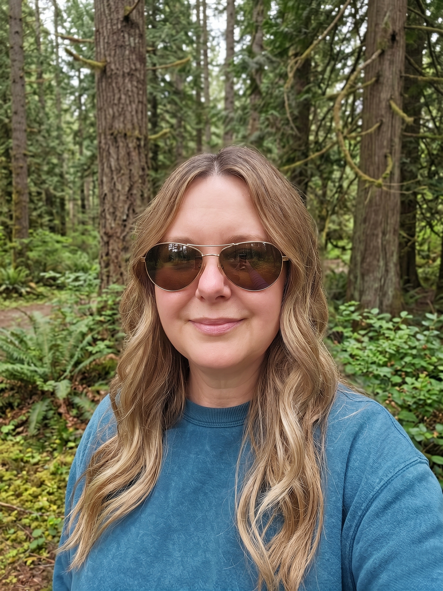 A middle aged woman wearing aviator sunglasses poses for a photo in a Pacific Northwest forest.