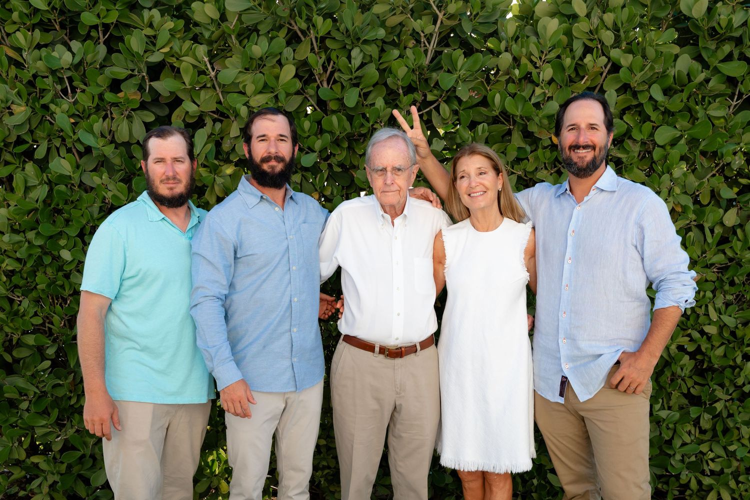 Family of five smiling together outdoors in front of lush green foliage, dressed in casual summer attire.