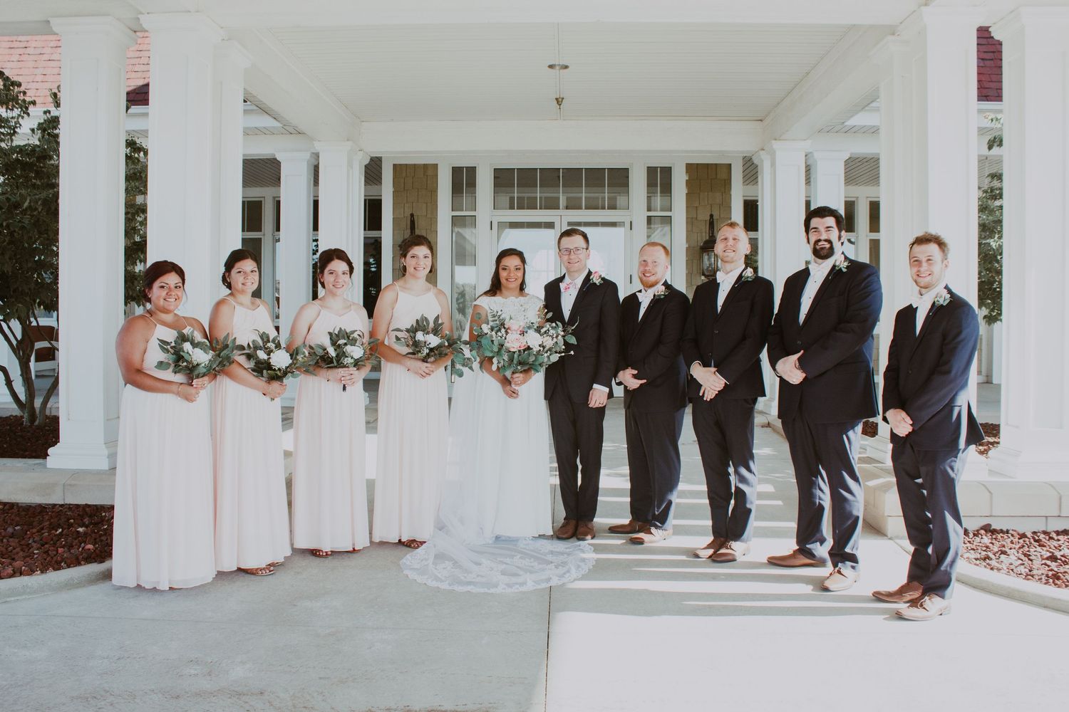 Wedding party posing in blush dresses and black suits on steps of a white columned venue.
