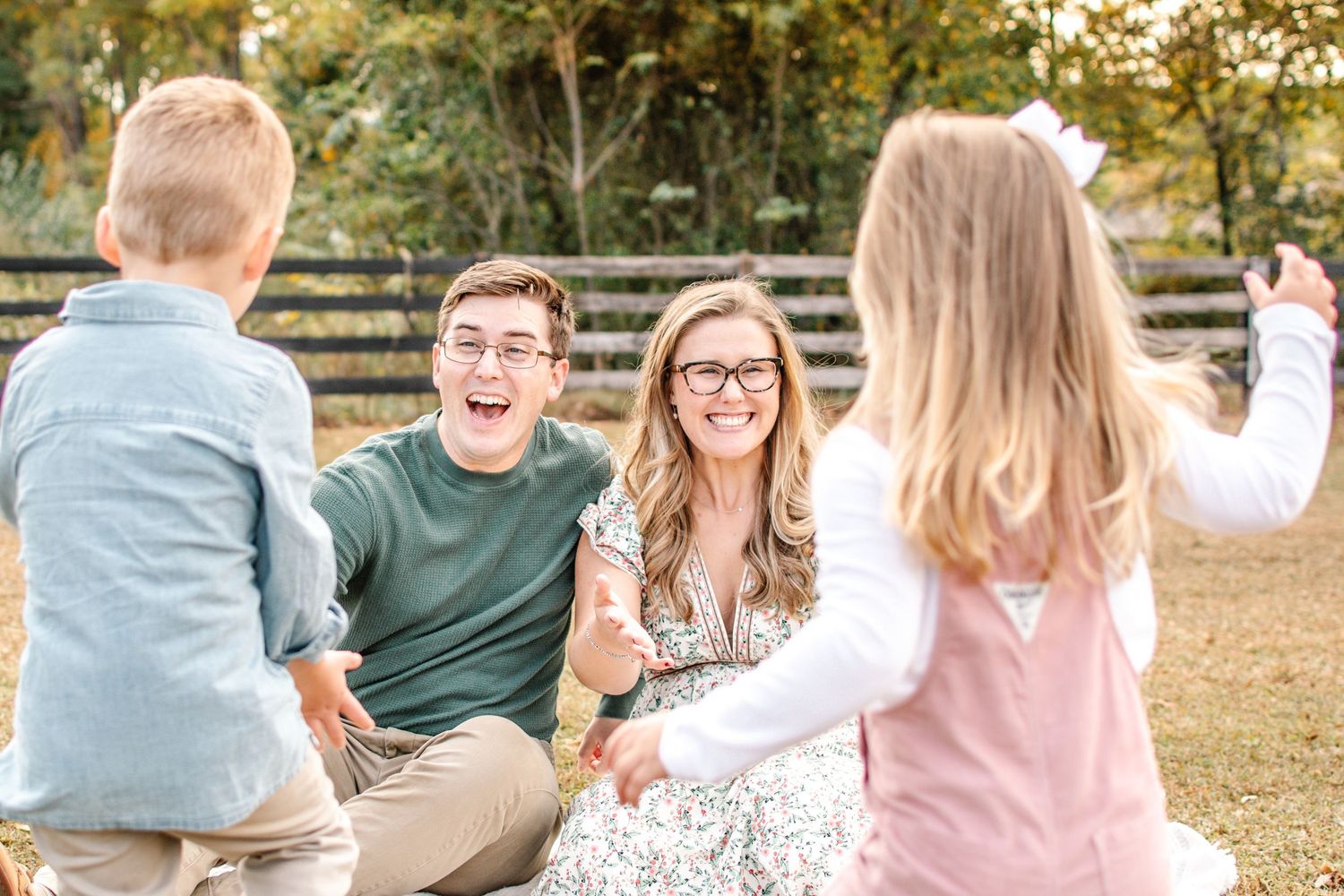 A joyful family sits on grass outdoors, laughing and playing with two young children during a sunny fall photo session.