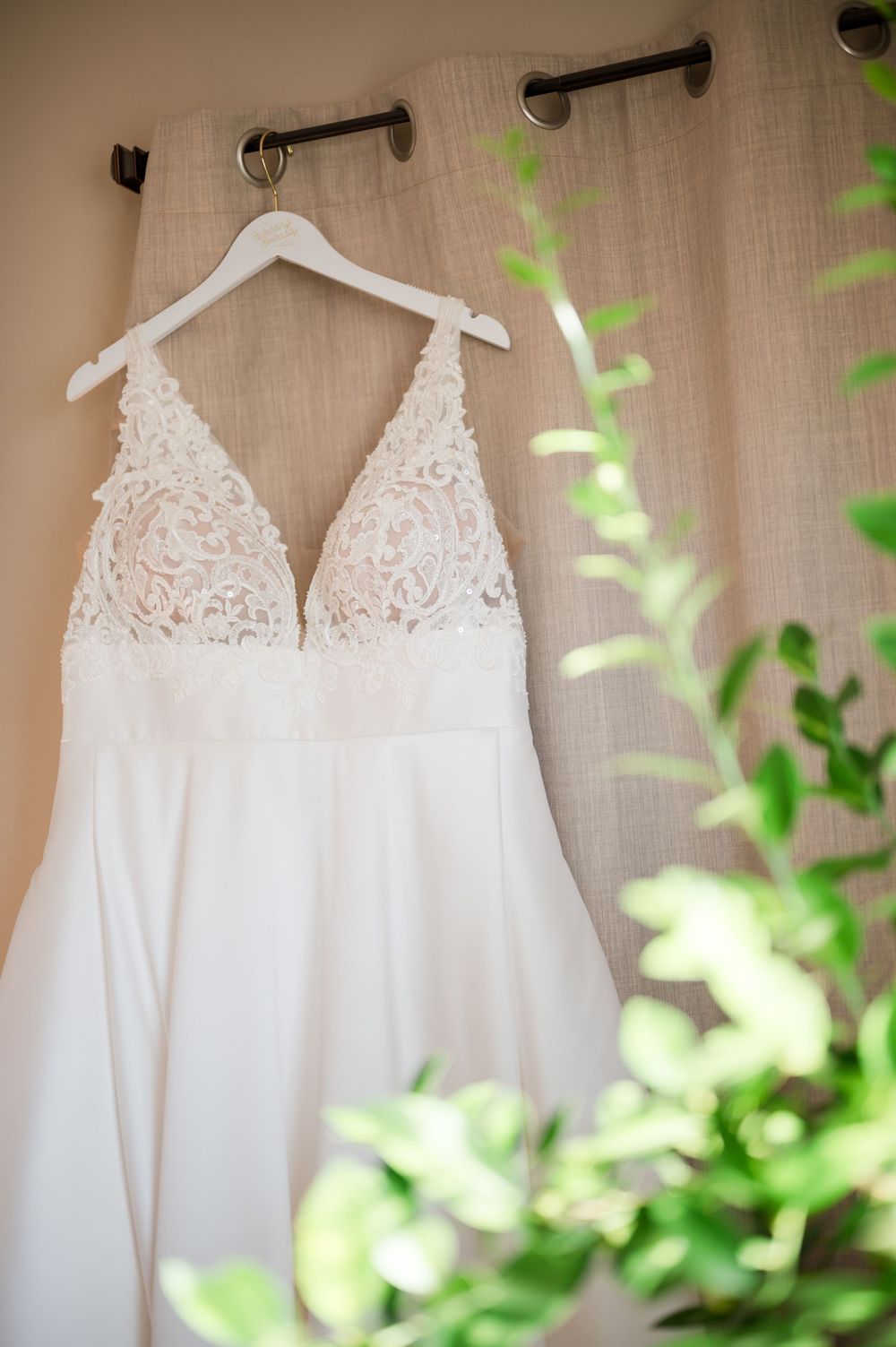 White lace bodice wedding dress hanging on a white hanger against neutral curtains with green foliage in foreground.