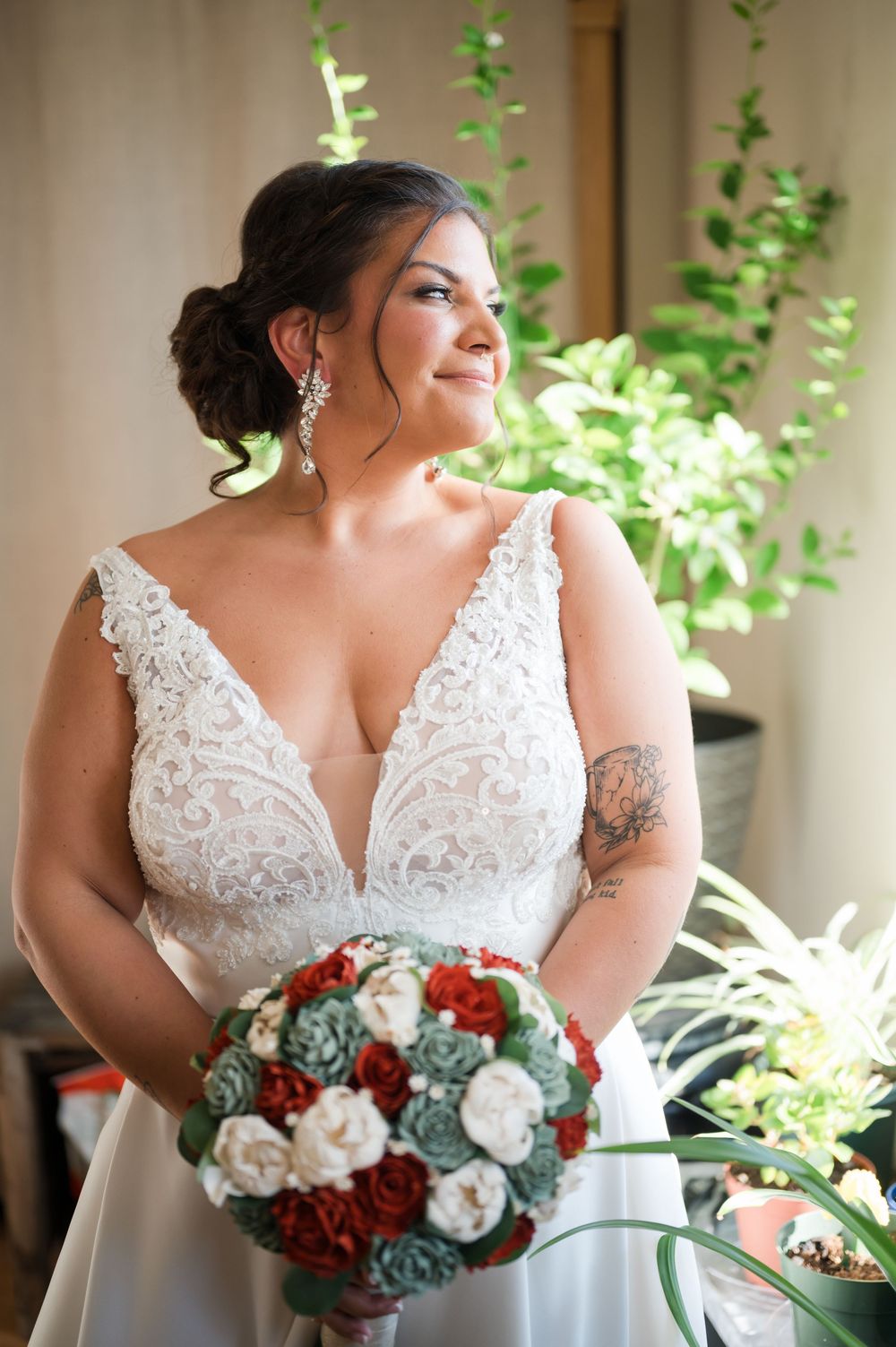 Bride in white lace V-neck gown holds red and white bouquet, smiling softly near greenery in natural light.