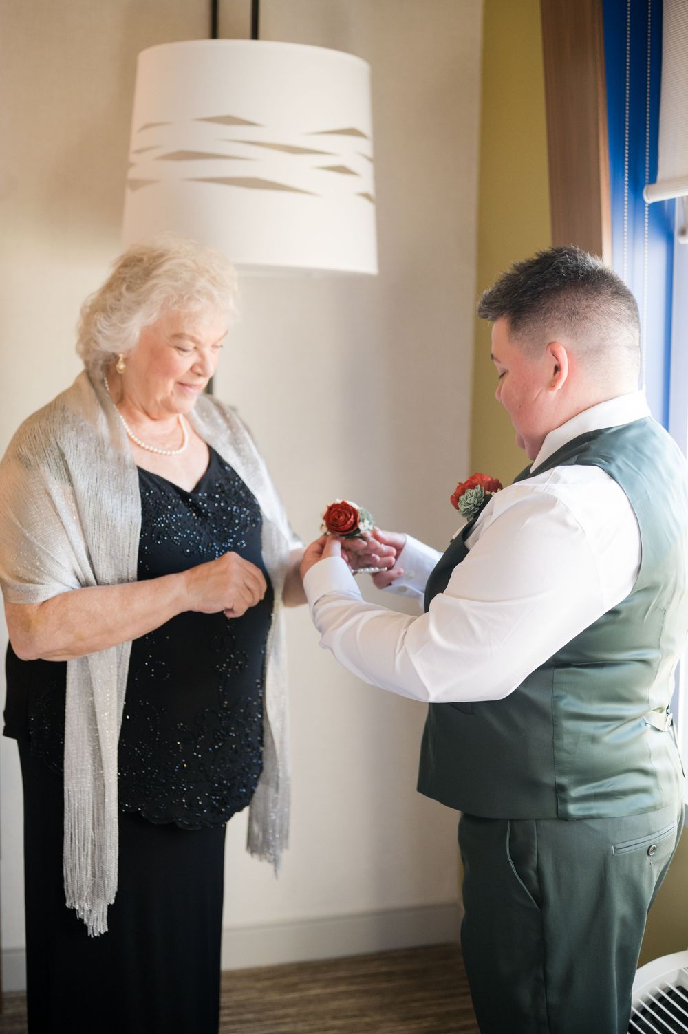 Groom in green vest pins red boutonniere on elderly woman in black sequin dress at wedding.