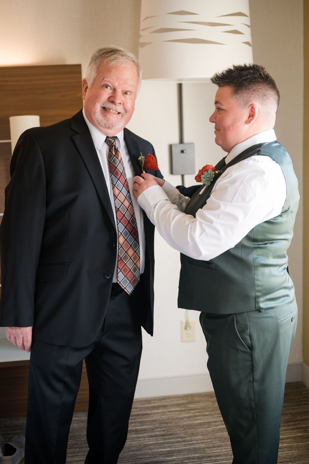 A groom in a green vest pins a boutonniere on an older man in a black suit during wedding preparations.