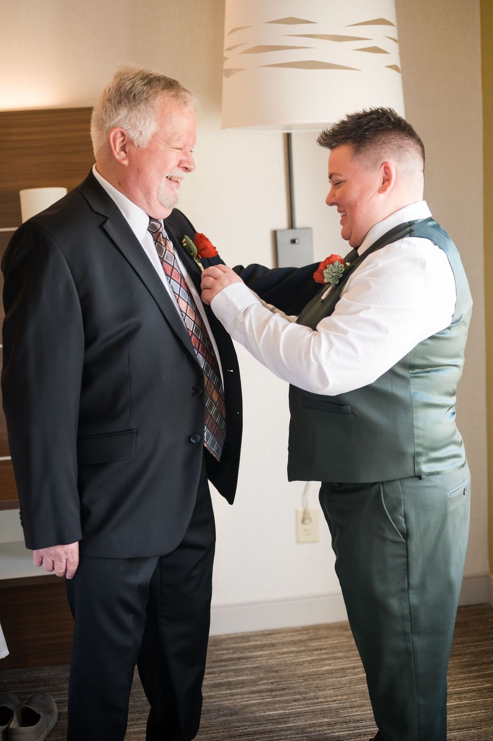 Groom pins boutonniere on older man in dark suit during wedding preparations in hotel room.