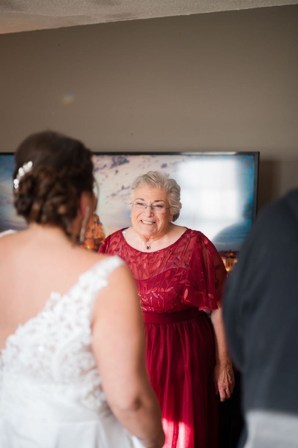An older woman in a red dress smiles warmly at a bride in a white lace gown before the wedding ceremony.