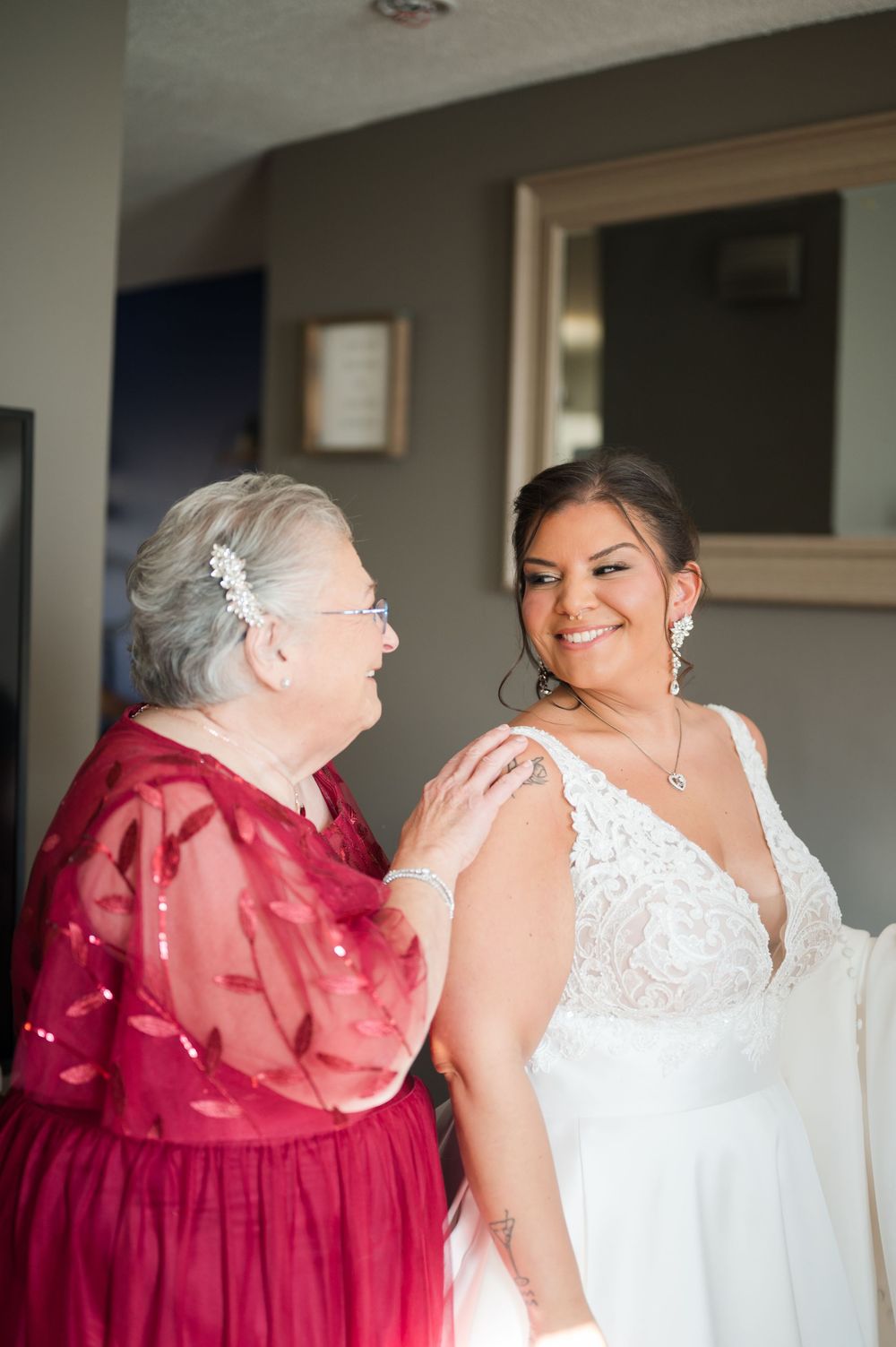 Elderly woman helping bride in lace wedding gown get ready, sharing a joyful moment before the ceremony.