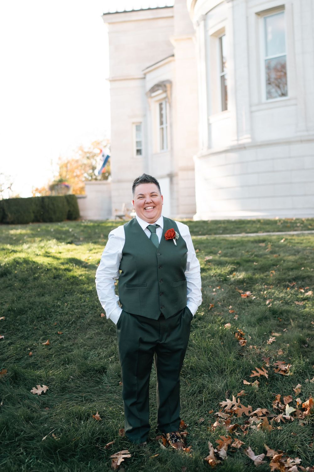 Person in green suit with red boutonniere smiling outdoors near elegant building on autumn day.