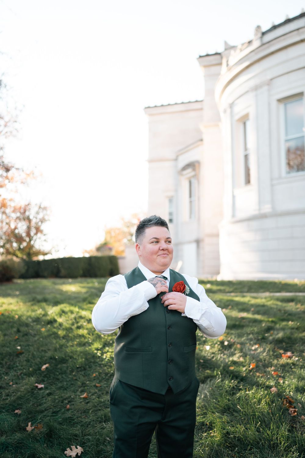 Person in green vest and tie adjusting boutonniere outdoors near elegant white building on wedding day.