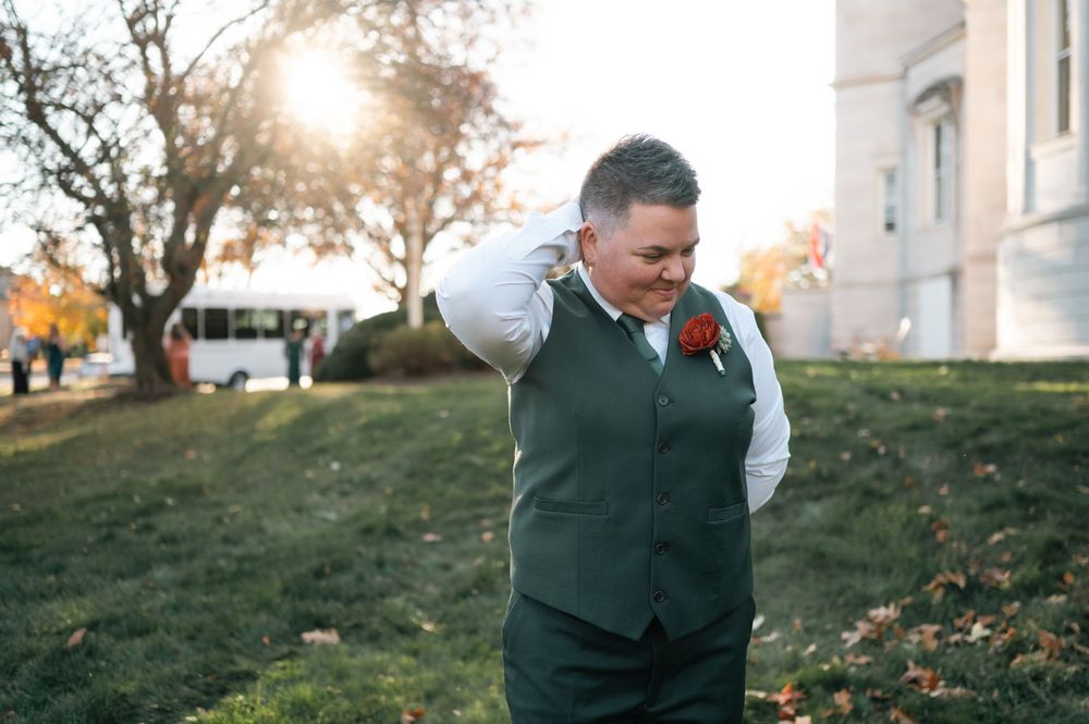 Groom in green vest and red boutonniere smiling outdoors at golden hour during fall wedding ceremony.