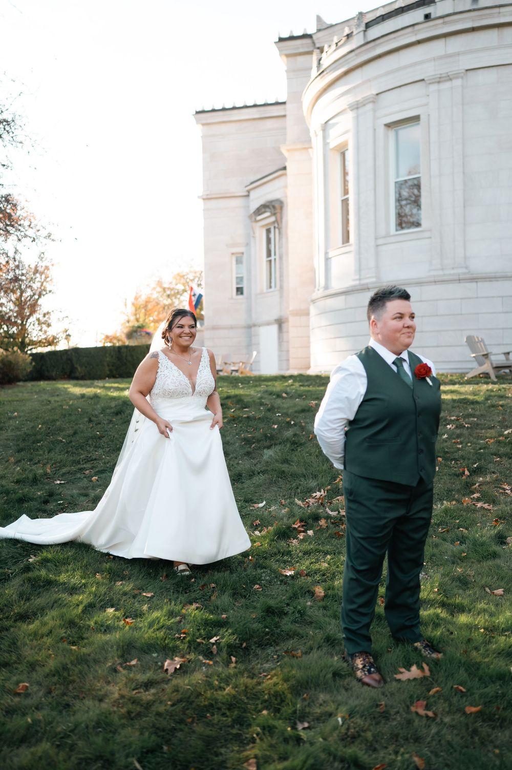 Bride in white gown approaches groom in dark green suit during first look at elegant stone mansion.