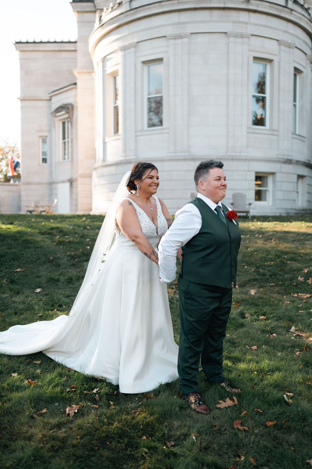 Two brides pose together outside a historic stone building, one in a white gown with veil and one in a green vest.