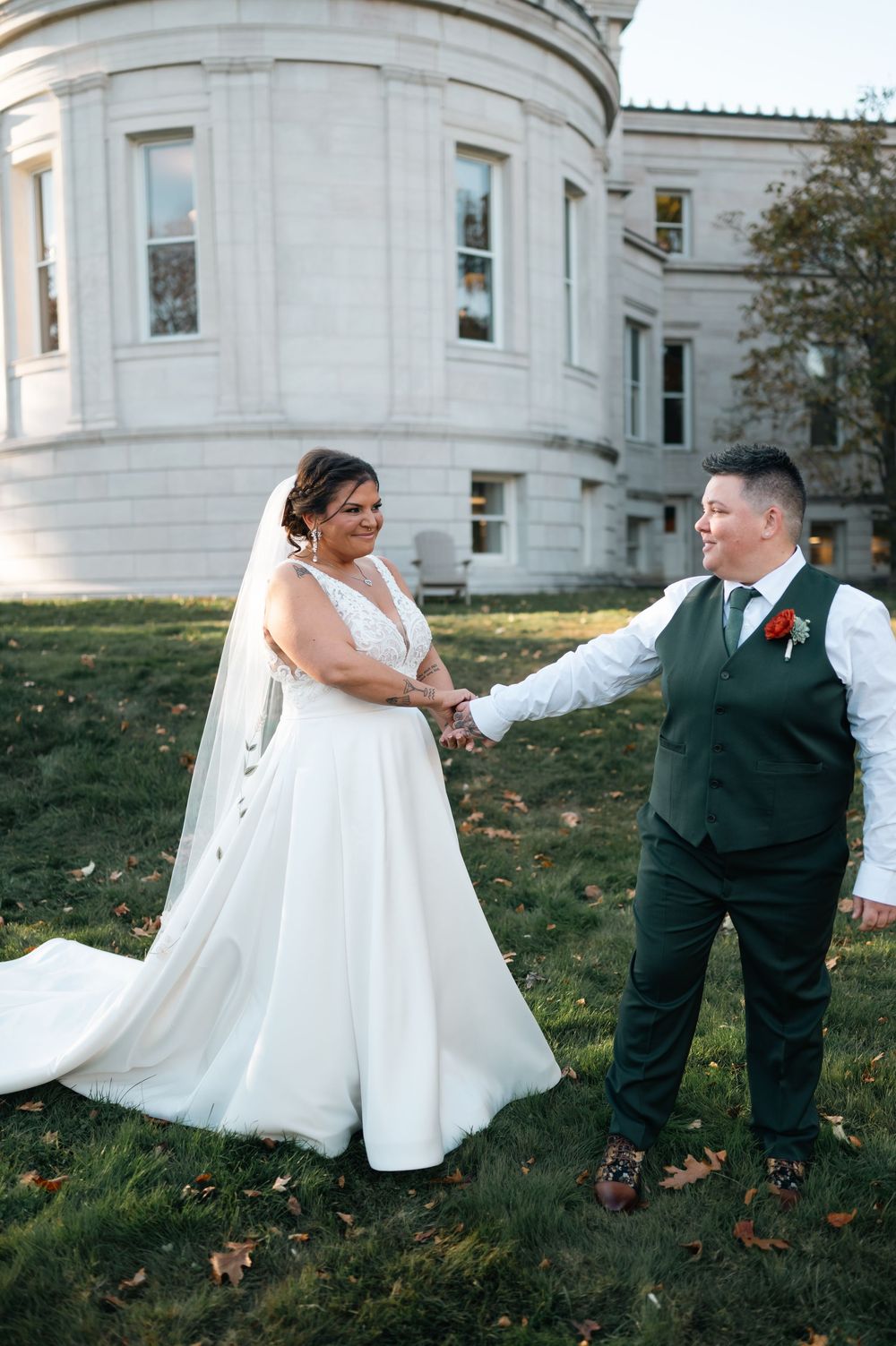 Two brides hold hands smiling in front of neoclassical building during outdoor wedding portraits.