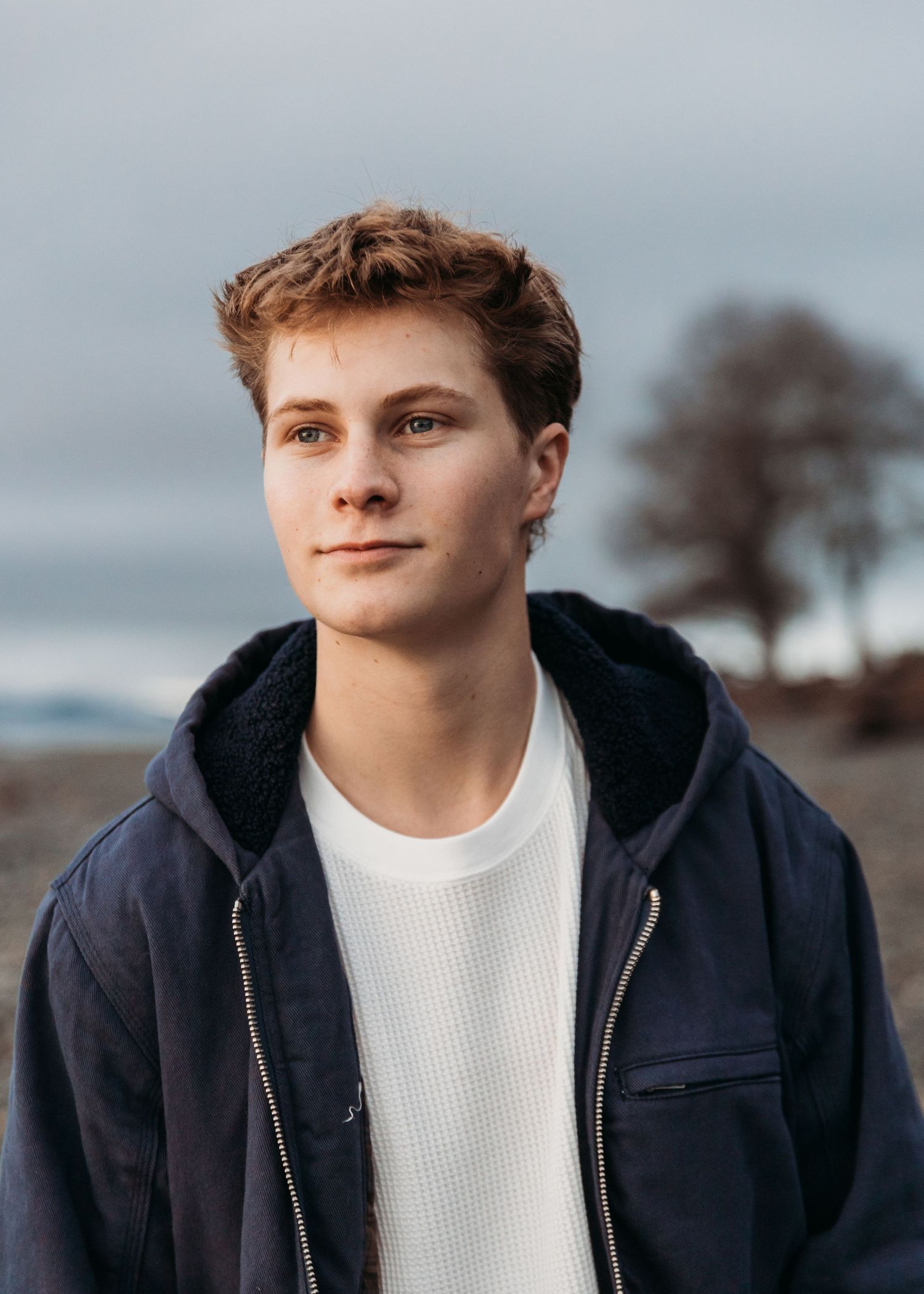 Young man in navy jacket and white sweater poses outdoors near beach with overcast sky.