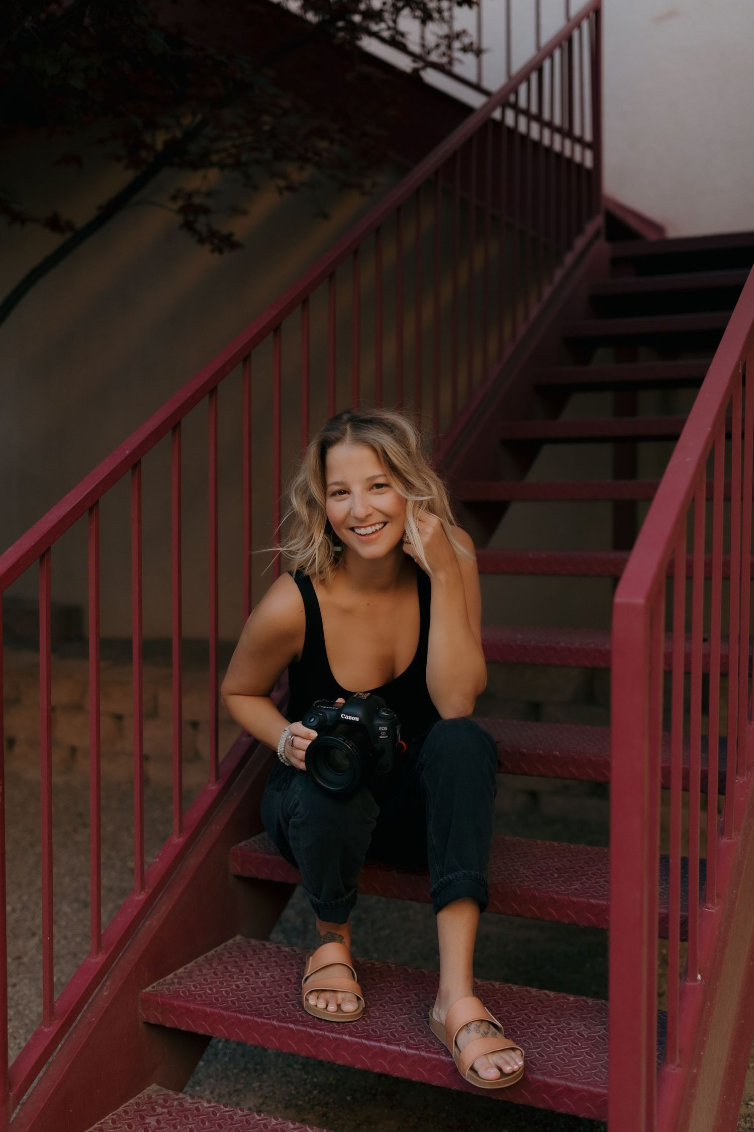 Young woman in black tank top and jeans holding a tan wide-brim hat, wearing layered bracelets and rings.