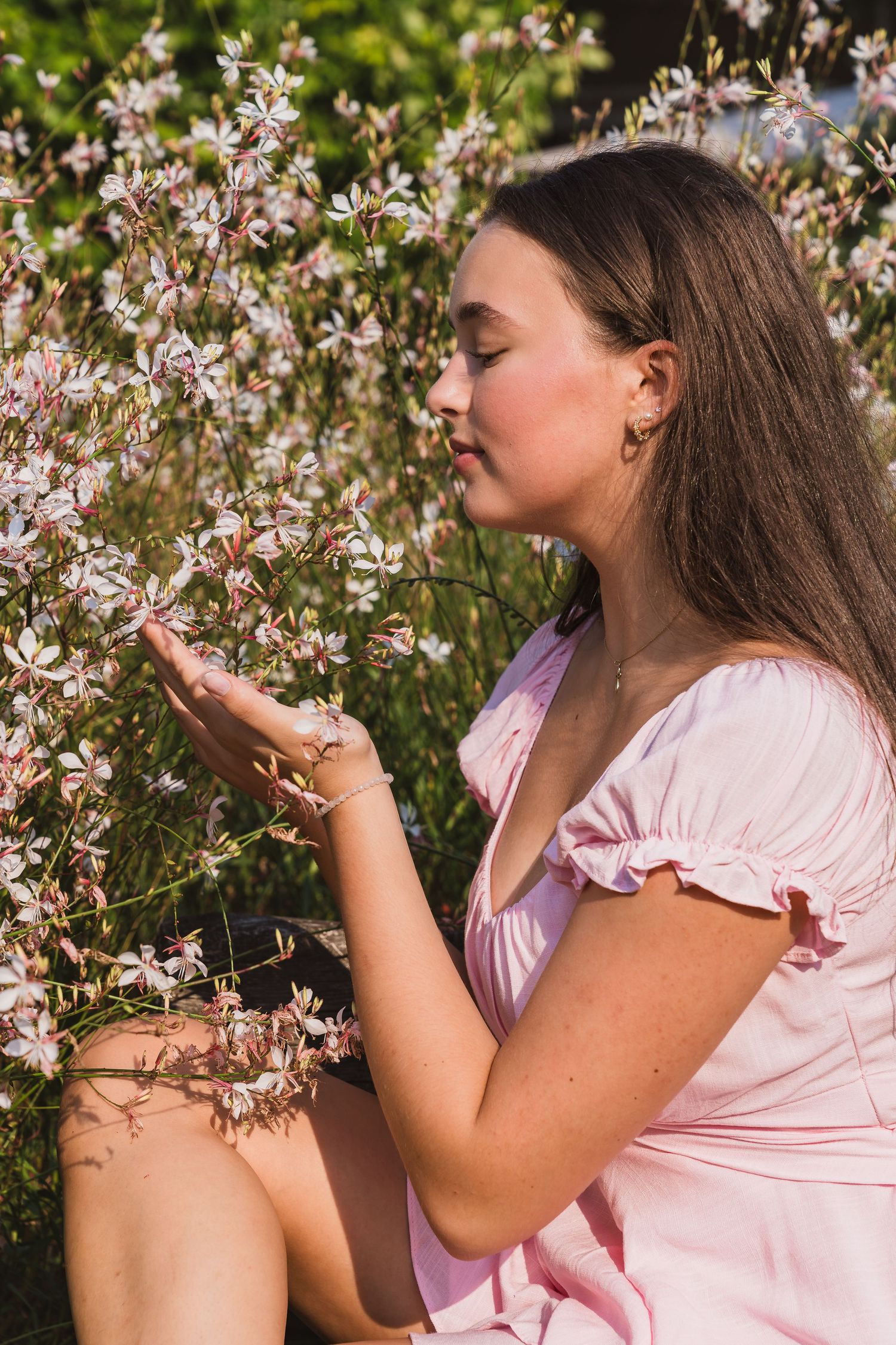 Young woman in pink dress smelling delicate white flowers outdoors on a sunny day.