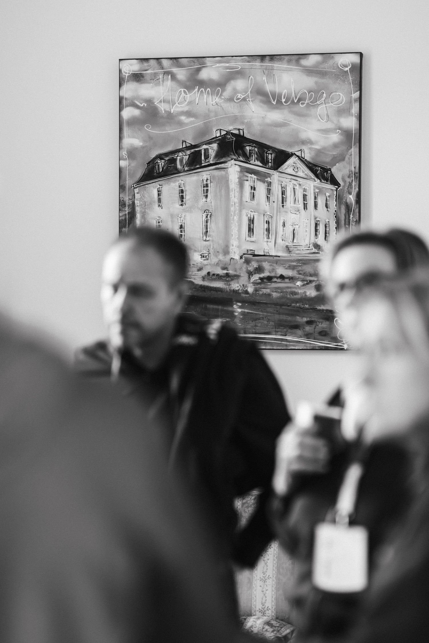 Black and white photo of people at an art gallery with a painting of a historic mansion on the wall.