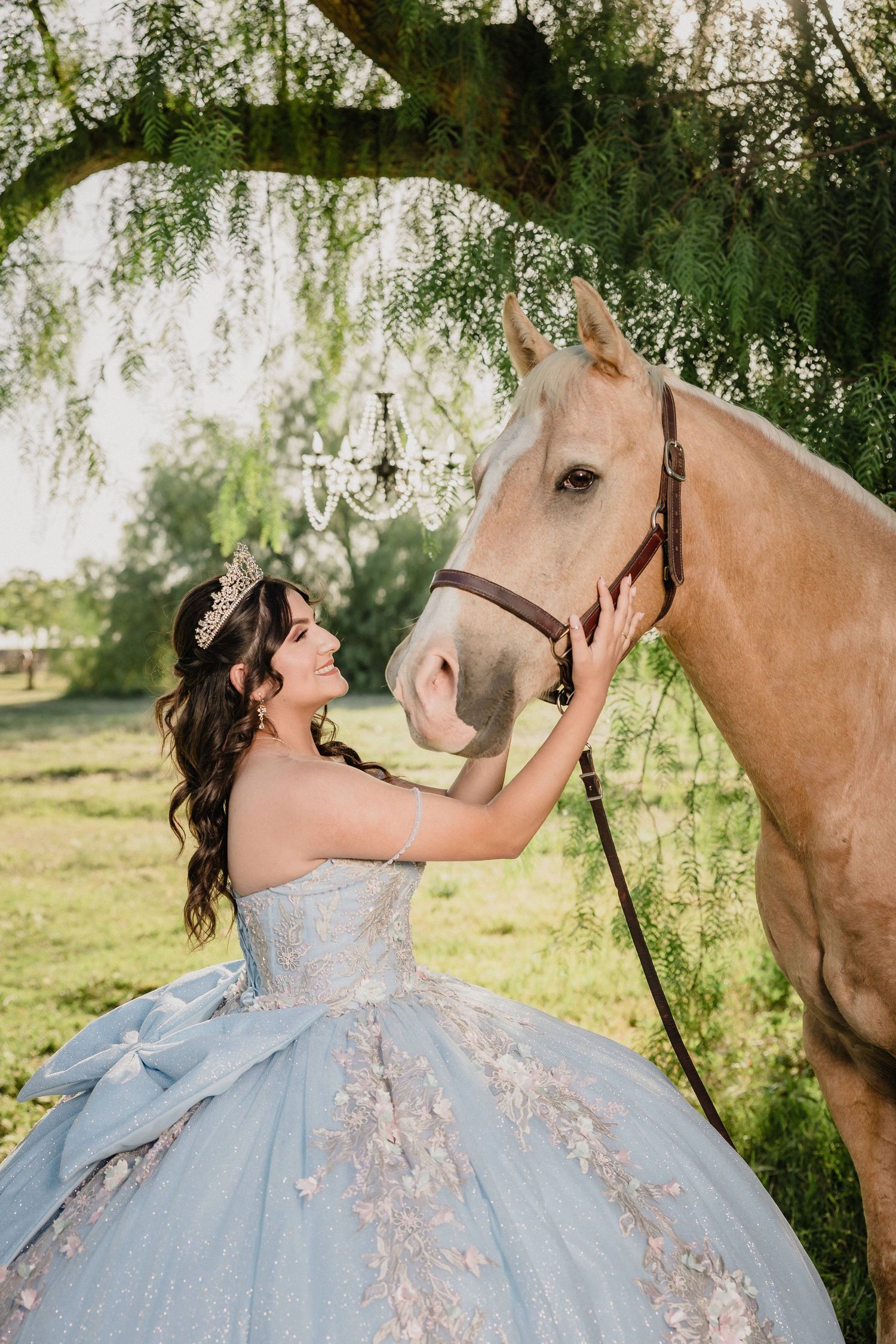 A young woman in a blue quinceañera gown and tiara gently touches a palomino horse under a willow tree.
