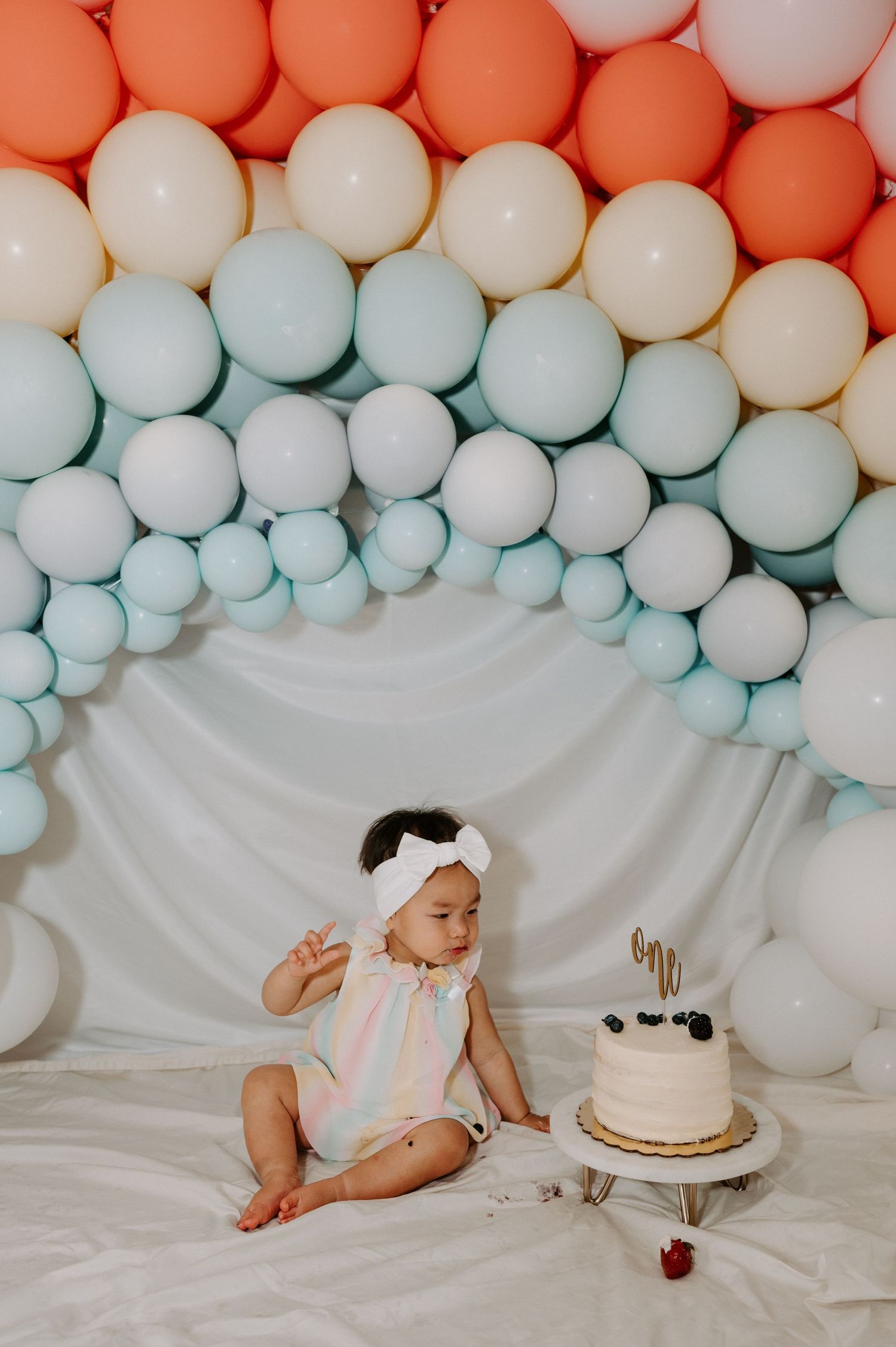 Baby girl in white romper smashes first birthday cake surrounded by colorful balloon arch backdrop.