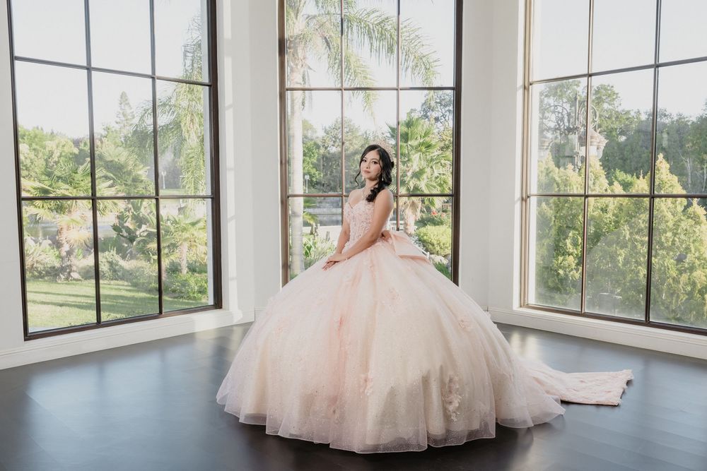 Young woman in a blush pink ball gown quinceañera dress poses in elegant room with large windows.