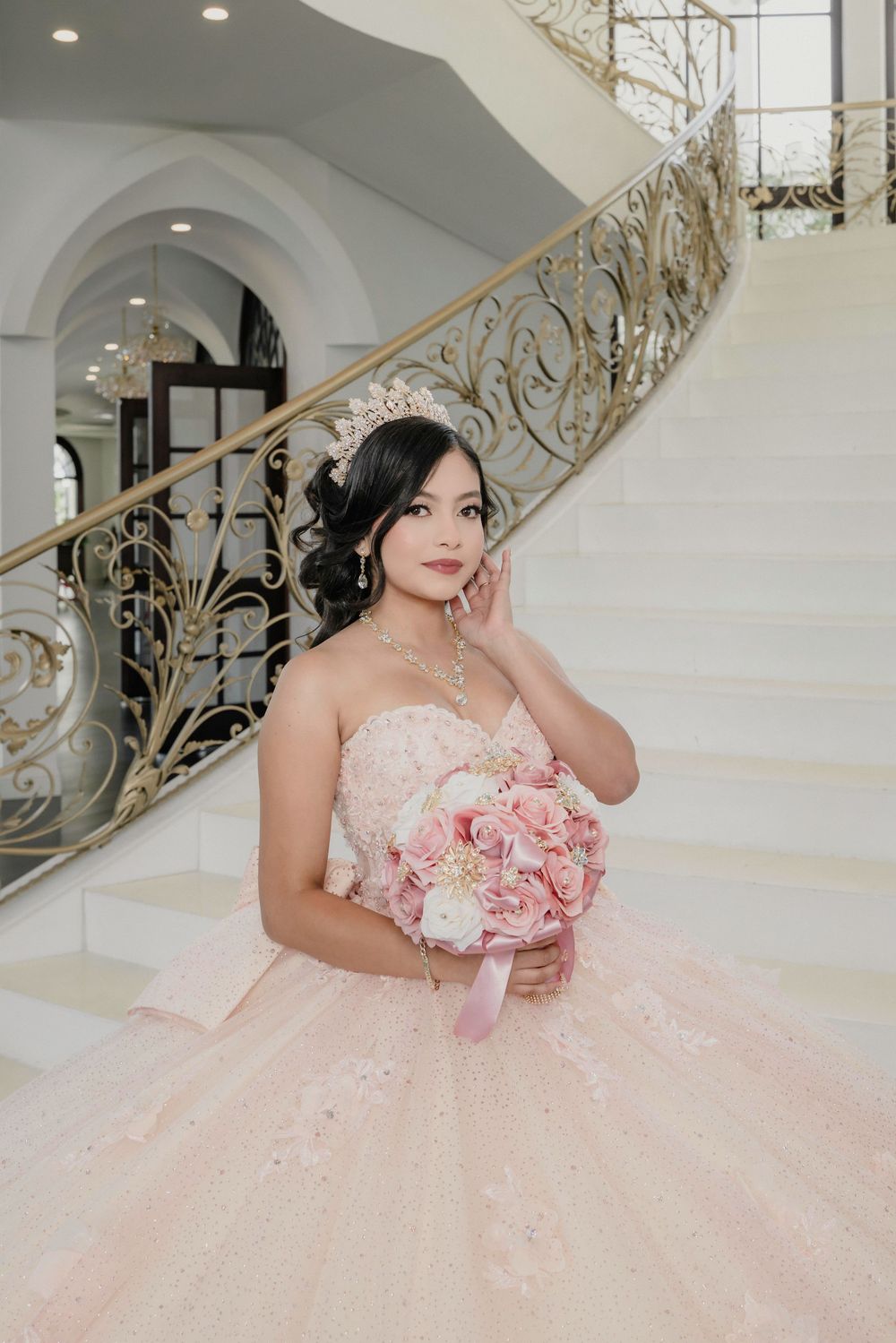 Young woman in pink ball gown with tiara holds floral bouquet on elegant staircase during quinceañera celebration.