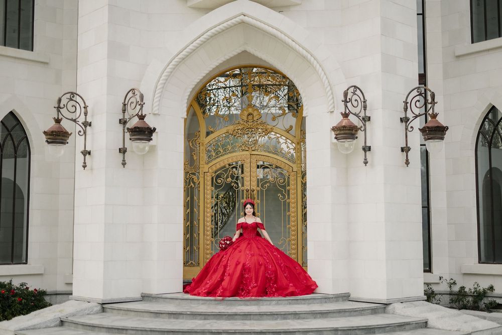 Woman in red ball gown posing at ornate golden gates of a white castle-like building on stone steps.