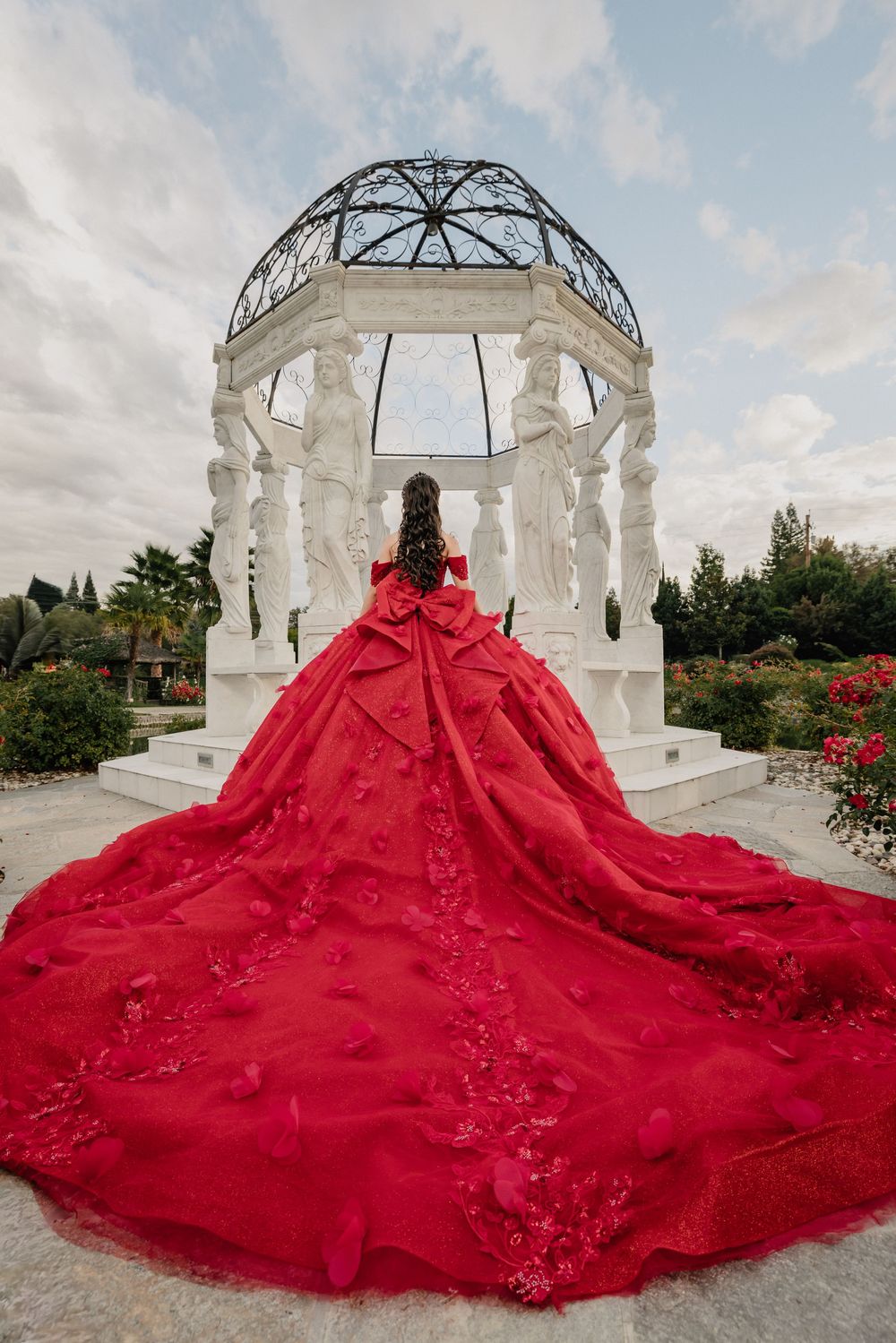Woman in stunning red ball gown with long train stands before elegant white gazebo in lush garden.
