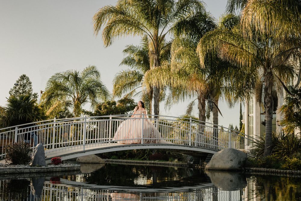 Bride in a flowing gown poses on a white bridge surrounded by palm trees, reflected in calm water at sunset.
