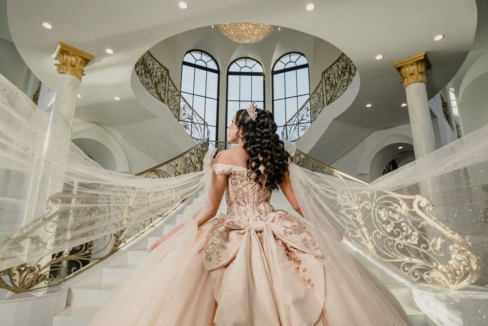 Bride in elegant champagne ball gown with gold embroidery and flowing veil stands on grand staircase in luxurious mansion.